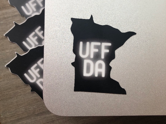 mage: Black die-cut sticker in the shape of the U.S. state of Minnesota. The word “UFFDA” is in the center of the sticker in glowing, white lettering